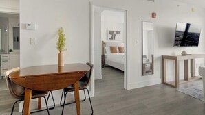 A modern hotel room with a wooden dining table, a vase with flowers, a bed with white bedding, a large mirror, and a TV on the wall. - Landing | Stylish 1BD, Gym (Hialeah)