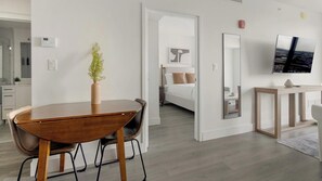 A modern hotel room with a wooden dining table, a vase with flowers, a bed with white bedding, a large mirror, and a TV on the wall. - Landing | Stylish 1BD, Gym (Hialeah)