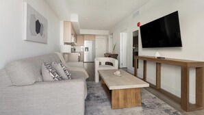A modern living room with a sofa, coffee table, TV, and kitchen in the background. - Landing | Stylish 1BD, Gym (Hialeah)