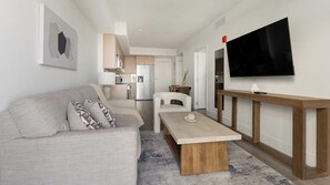 A modern living room with a sofa, coffee table, TV, and kitchen in the background. - Landing | Stylish 1BD, Gym (Hialeah)
