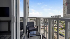 A balcony with a black chair, a TV on the left, and a view of a parking lot and buildings. - Landing | Stylish 1BD, Gym (Hialeah)