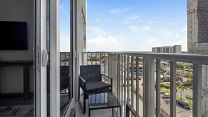 A balcony with a black chair, a TV on the left, and a view of a parking lot and buildings. - Landing | Stylish 1BD, Gym (Hialeah)