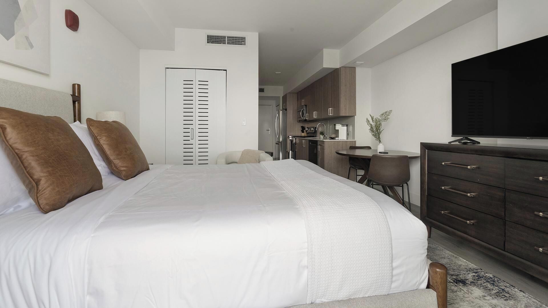 Een moderne slaapkamer met een groot bed, een dressoir, een televisie en een kitchenette op de achtergrond.