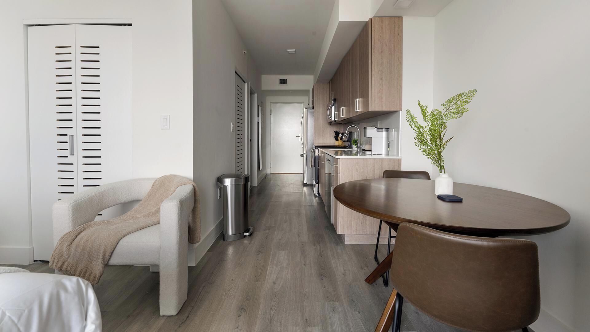 Een moderne keuken met roestvrijstalen apparatuur, een eethoek met een ronde tafel en stoelen, en een gang die naar een slaapkamer leidt.
