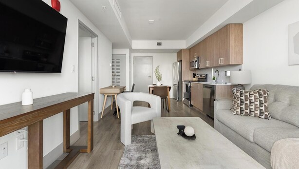 A modern living room with a flat-screen TV, a dining area, a kitchen with wooden cabinets, and a grey sofa. - Landing | Stunning 1BD, Gym (Hialeah)