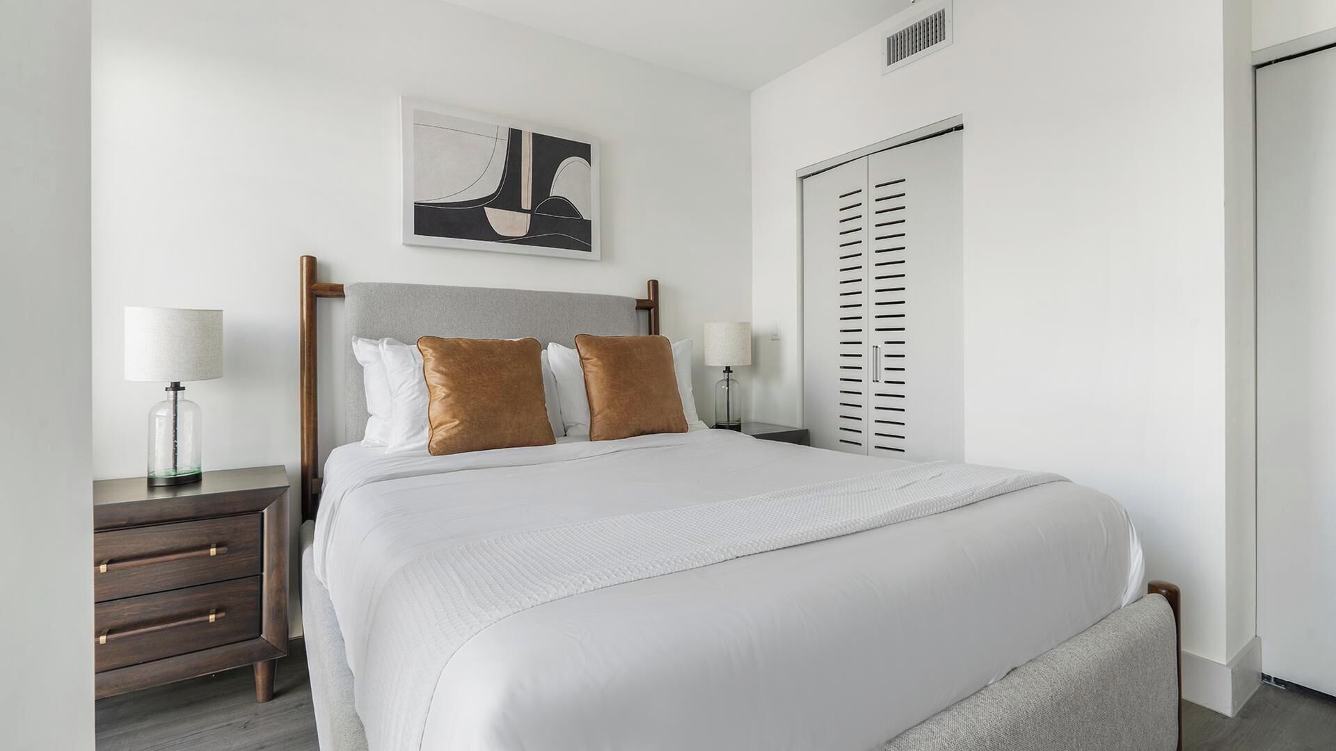 Habitación de hotel ordenada con cama, mesitas de noche, un armario y una obra de arte colgada en la pared.