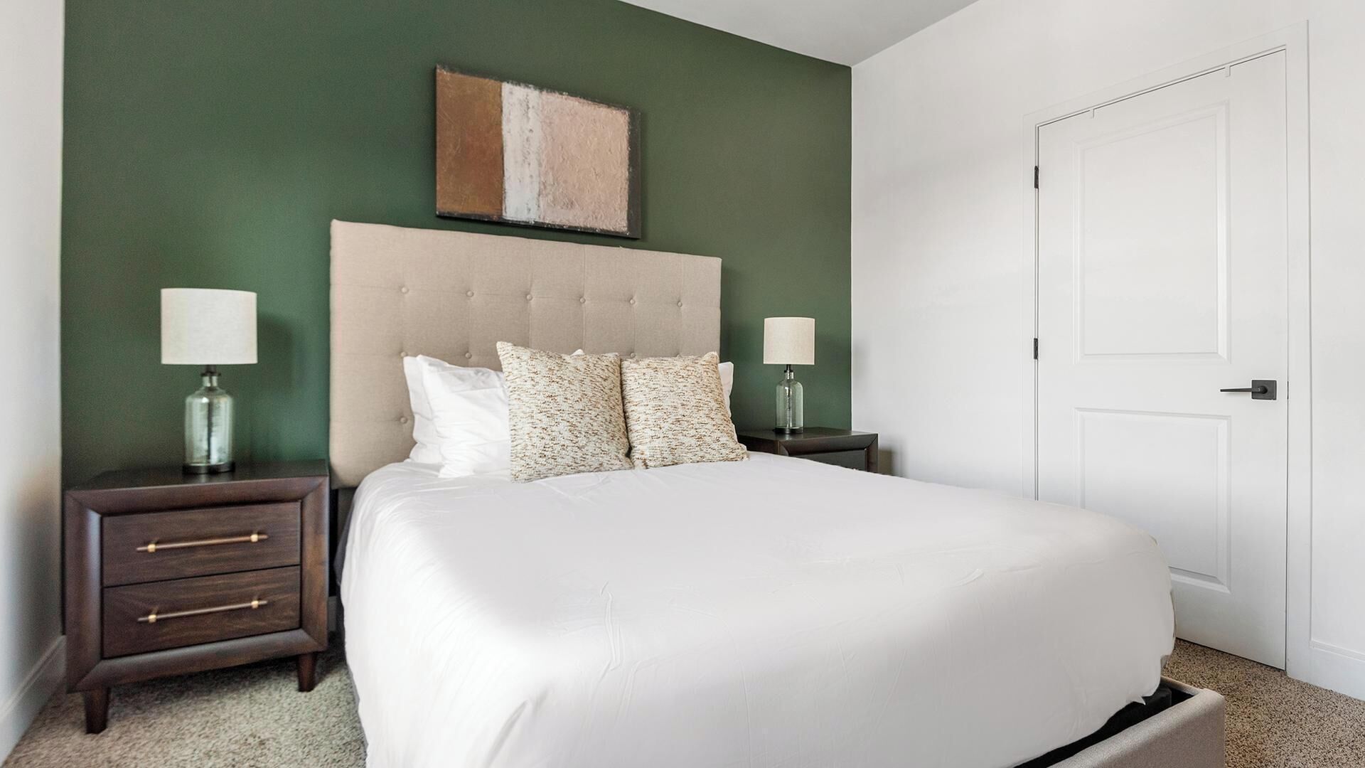 Un dormitorio con una cama grande, dos mesitas de noche con lámparas, una mesita de madera y una puerta blanca, además de un muro verde decorativo.