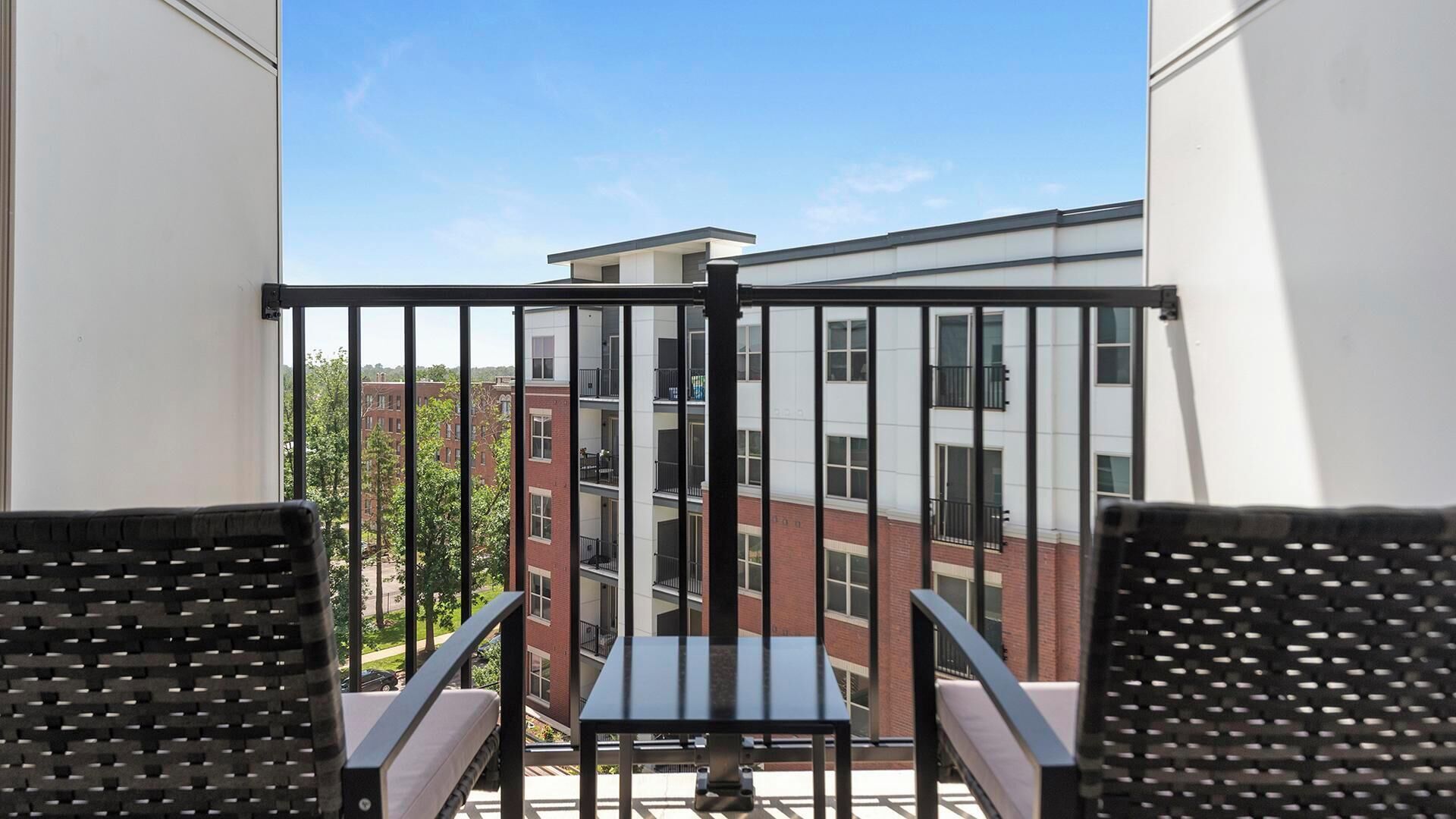 A balcony with two chairs and a small table, overlooking a building with multiple windows and a brick facade.