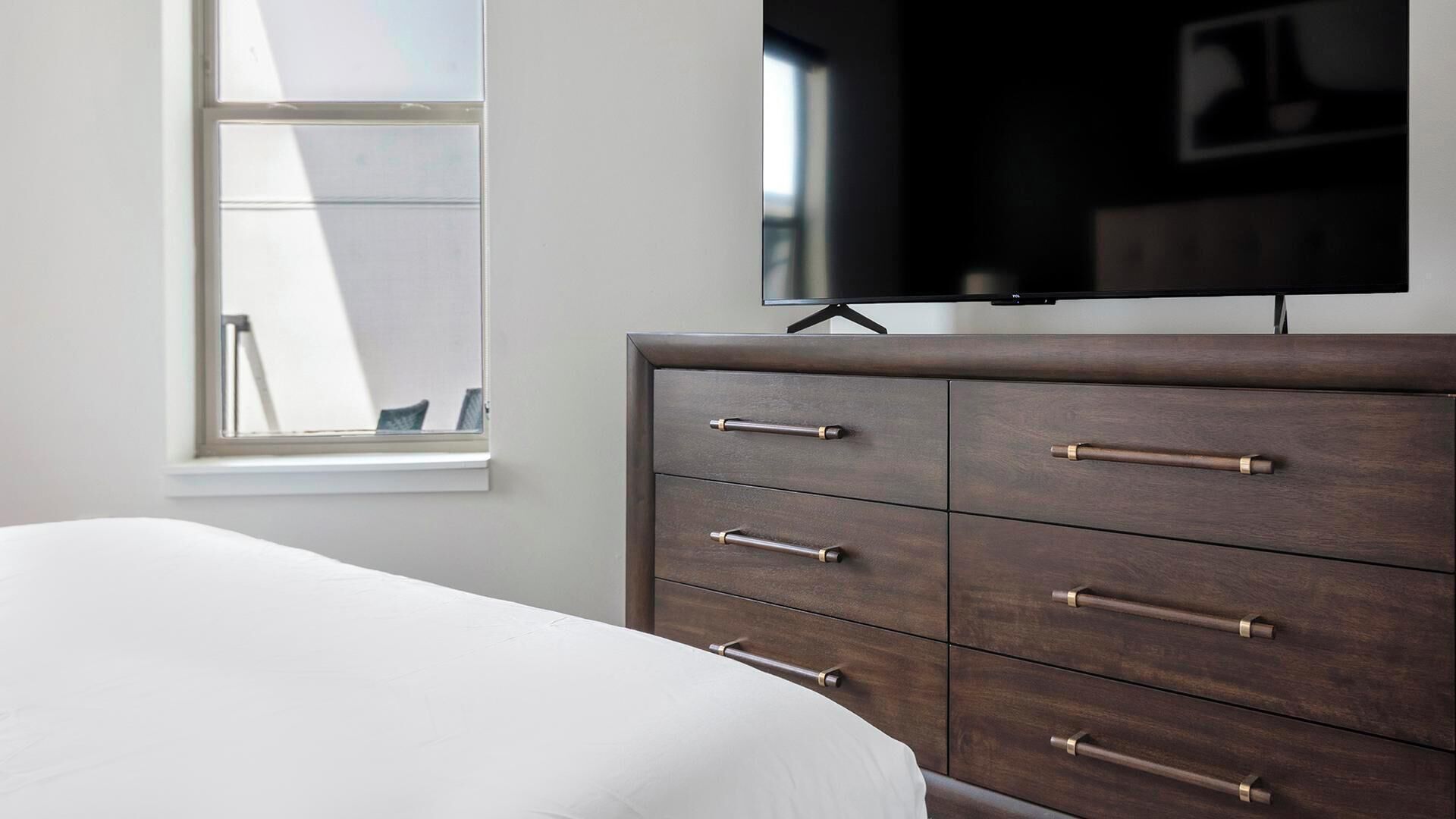 A bedroom with a bed, a dresser with drawers, a television on a stand, and a window.
