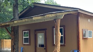 A small wooden cabin with a porch, two windows, a door, and a small air conditioning unit. - Whoa-Man Ultra Cozy Aggieland  Cabin (North Zulch)