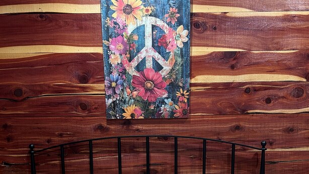 A bed with a yellow floral bedspread and matching pillows, a metal headboard, and a wooden wall with a peace sign painting. - Whoa-Man Ultra Cozy Aggieland  Cabin (North Zulch)