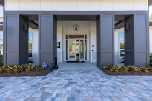 A modern building entrance with a glass door, a sign indicating a no-smoking area, and a paved walkway with plants. - Landing | Stunning 2BD, Clubhouse (Pensacola)