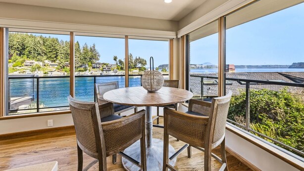 A dining area with a round table and four chairs, large windows offering a view of a body of water and a hillside. - Luxury Waterfront Home walk to Downtown Gig Harbor (Gig Harbor)
