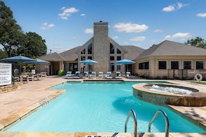 A swimming pool with lounge chairs and umbrellas, a stone building with large windows, and a fenced area. - Landing | Stunning 2BD, Gym (Lewisville)