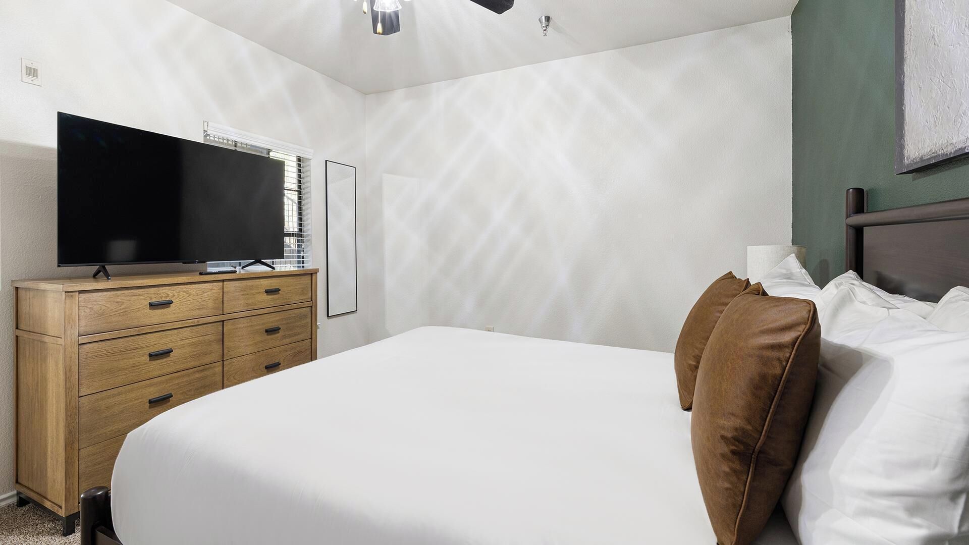 A bedroom with a bed, a wooden dresser, a flat-screen TV, and a wall-mounted light fixture.