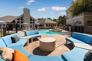 A pool area with blue outdoor seating and a stone building in the background. - Landing | Incredible 2BD, Gym (Lewisville)