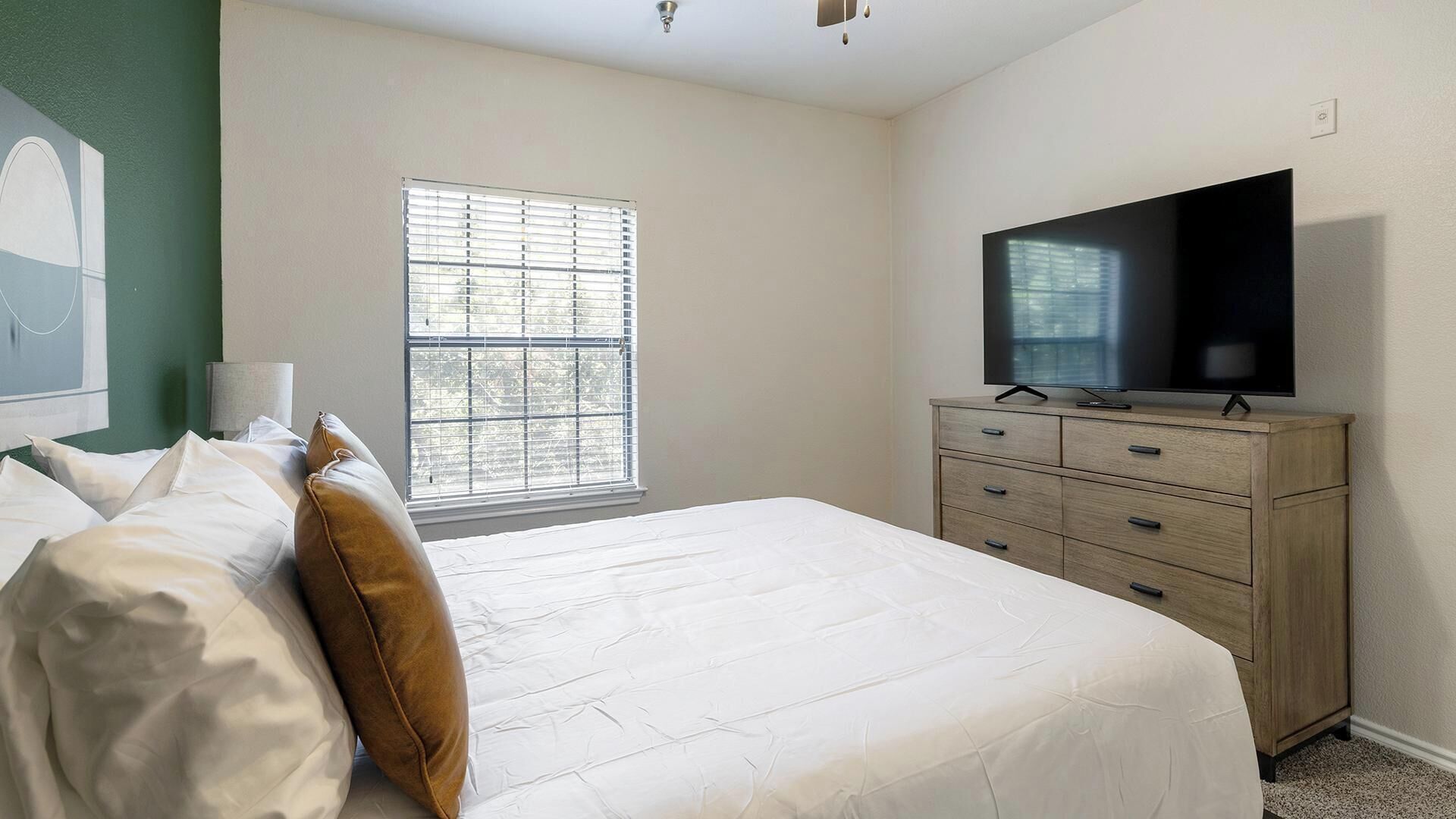 Une chambre à coucher comprenant un lit, une commode, un téléviseur et une fenêtre avec des stores.