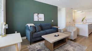 A living room with a sofa, a coffee table, and a dining table. There is a kitchen area with a sink and cabinets. - Landing | Spectacular 2BD in Frisco - Fields (Frisco)