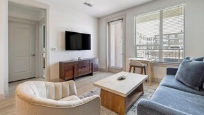 A modern living room with a flat-screen TV, a wooden coffee table, a beige sofa, a gray sofa, and a dining area with a chair and table. - Landing | Modern 1BD, Rooftop Lounge, Gym (Frisco)