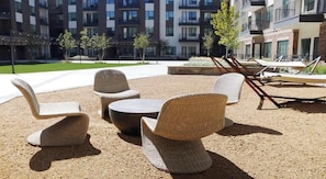Terrassenmöbel aus Rattan mit rundem Tisch, auf einem Kiesgrund in einem Innenhof, mit einem Gebäude im Hintergrund.