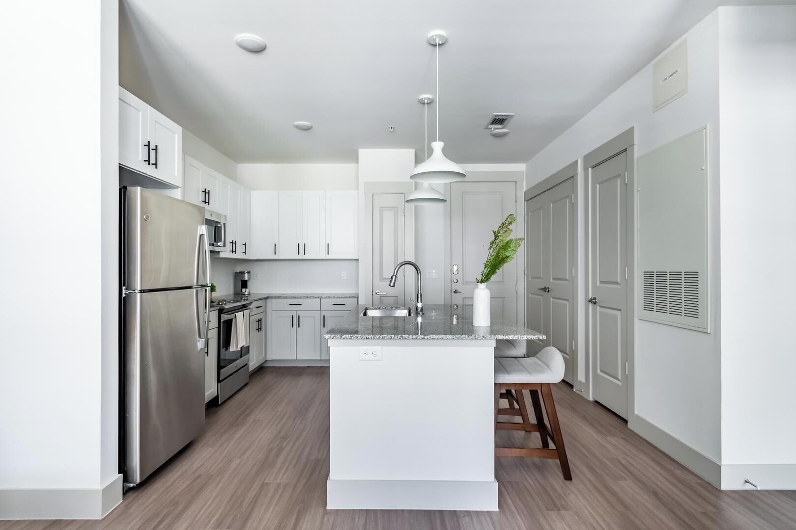 Une cuisine moderne avec des meubles blancs, des appareils en acier inoxydable, un îlot central doté d’un évier, des luminaires suspendus et une plante.
