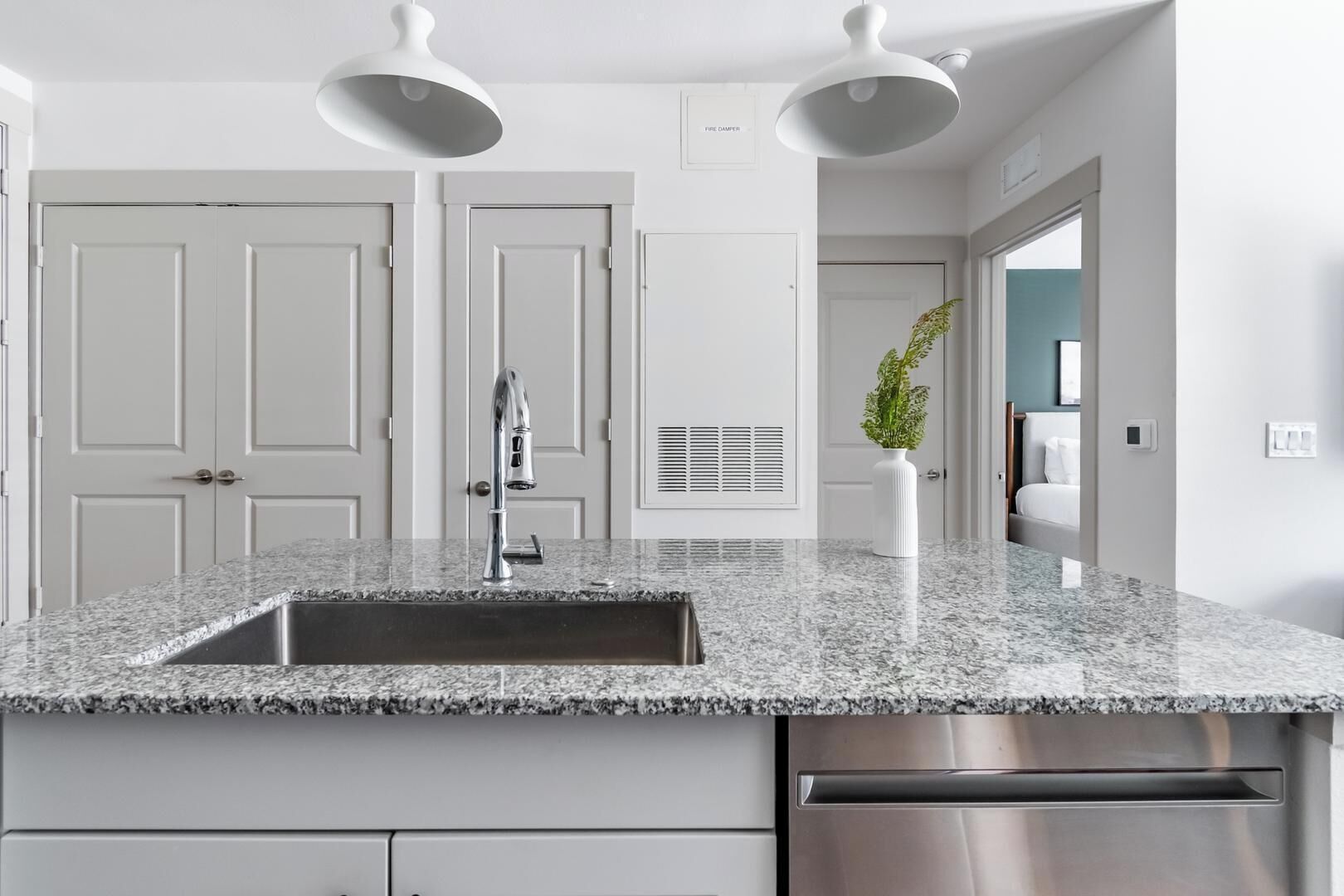 Una cocina con encimera de granito, pileta de acero inoxidable y lavavajillas. Se observan armarios y una puerta blancos al fondo.