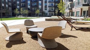 Terrassenmöbel aus Rattan mit rundem Tisch, auf einem Kiesgrund in einem Innenhof mit Blick auf moderne Apartmenthäuser.
