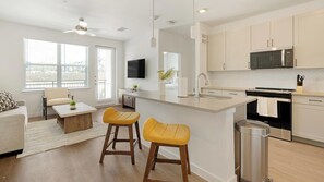 A modern kitchen with a central island, a dining area with a sofa and chairs, and a TV mounted on the wall. - Landing | Chic 1BD, Pool, Gym (Dallas)