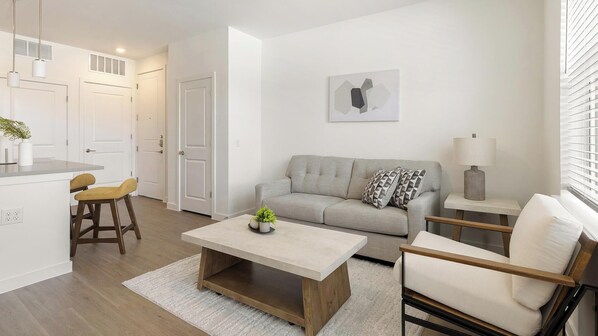 En moderne stue med sofa, sofabord og stole, et køkkenområde med en barstol samt et spisebord.