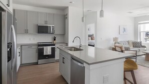A modern kitchen with stainless steel appliances, a central island, and a view into the living area. - Landing | Amazing 2BD, Courtyard, Gym (Dallas)