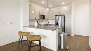 A modern kitchen with white cabinets, a stainless steel refrigerator, and a central island with stools. - Landing | Spectacular 1BD, Courtyard, Pool (Dallas)