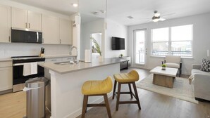 A modern kitchen with a central island, a dining area with a sofa and chairs, and a living room with a TV. - Landing | Chic 1BD, Pool, Gym (Dallas)