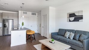 A modern kitchen with a central island, a grey sofa with patterned cushions, and a minimalist artwork on the wall. - Landing | Spectacular 1BD, Gym, Pool (Dallas)