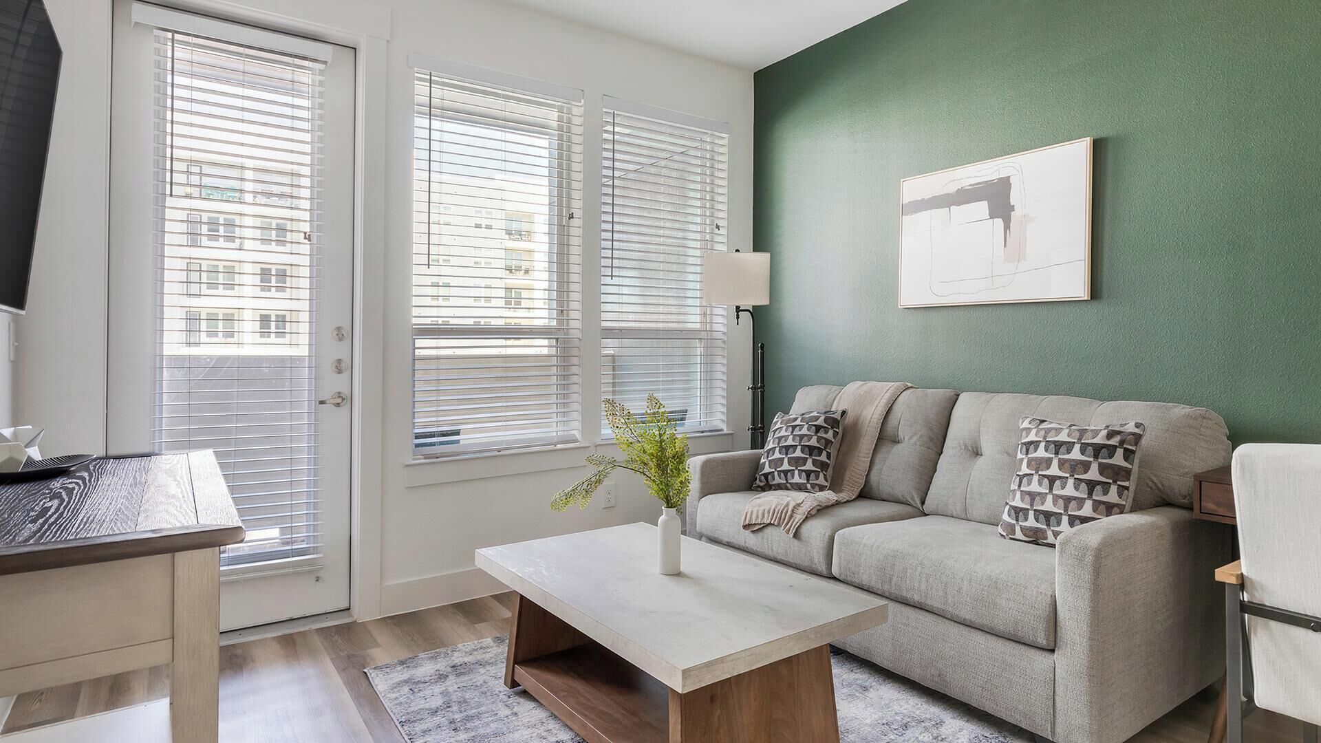Ein Wohnzimmer mit einer Couch, einem Couchtisch und einer Lampe. Eine Tür mit Jalousien und ein Fenster mit Jalousien sind vorhanden.