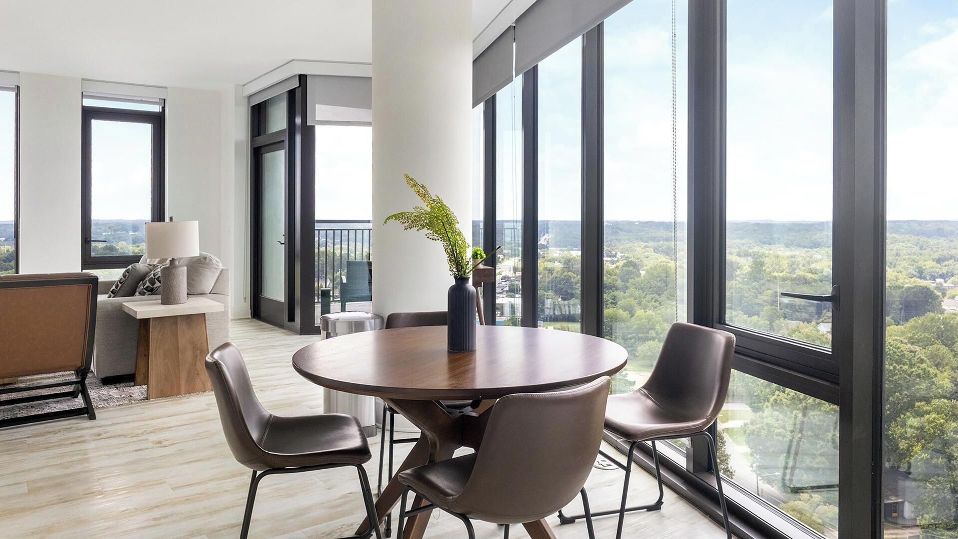 A modern room with a round table, two chairs, a sofa, and a view of a cityscape through large windows.