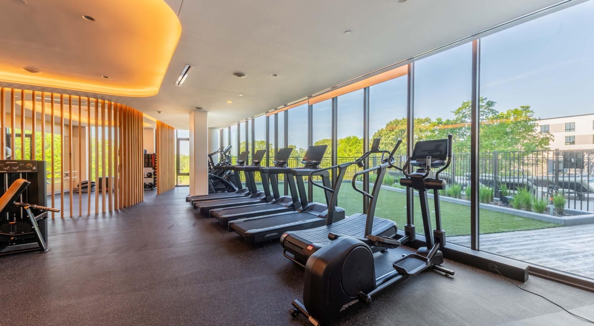 A modern gym with treadmills, a rowing machine, and a piano, featuring large windows and a wooden partition.