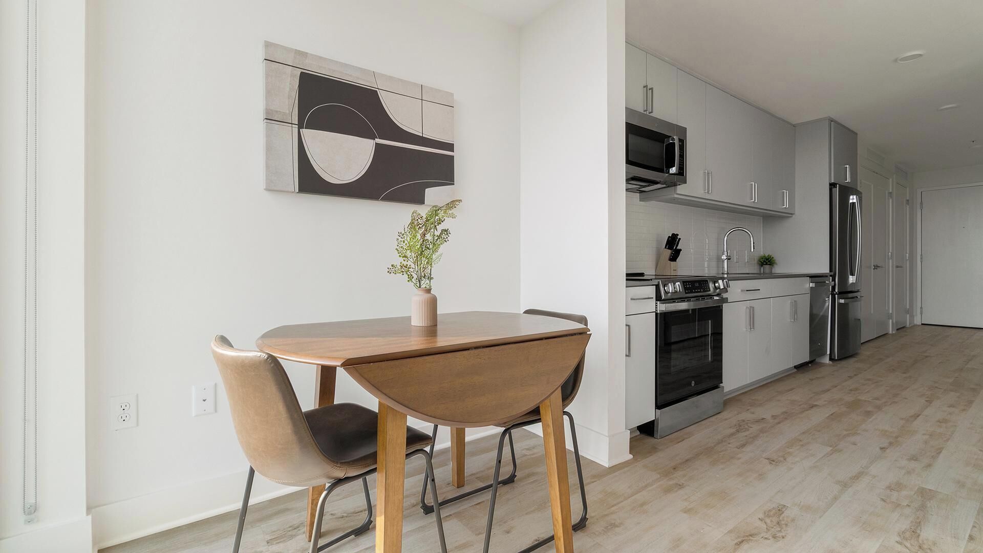 Eine moderne Küche mit Edelstahlgeräten, ein Essbereich mit einem Holztisch und zwei Stühlen sowie ein minimalistisches Wandbild.