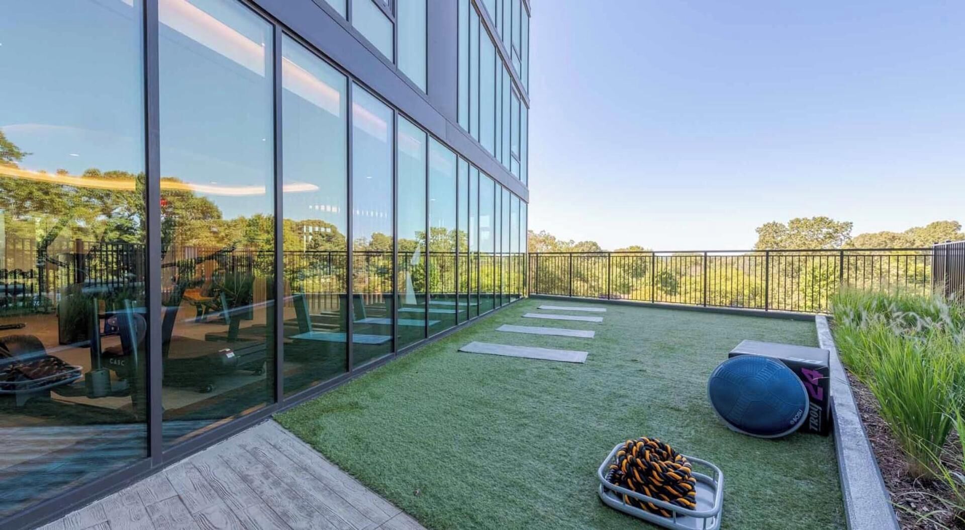 A modern building with large glass windows, a rooftop garden with artificial grass, outdoor seating, and exercise equipment.
