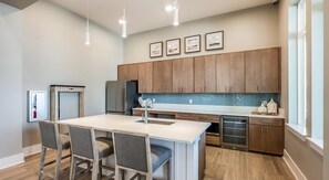 A modern kitchen with wooden cabinets, a central island with bar stools, stainless steel appliances, and a large window providing natural light. - Landing | Spectacular 3BD, Gym, Yoga Studio (Four Corners)