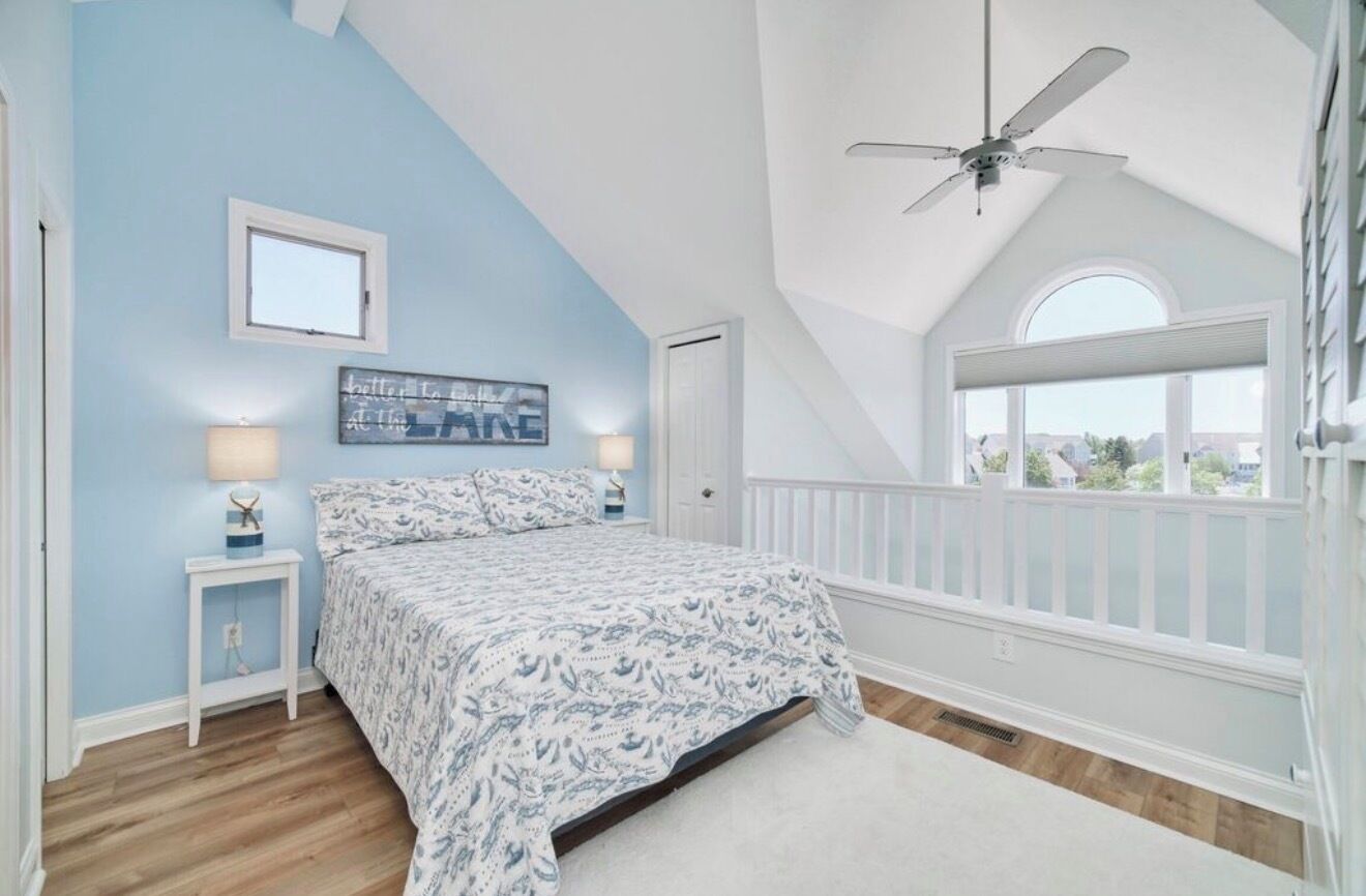 A bedroom with a sloped ceiling, a bed with patterned bedding, a nightstand with a lamp, a window with a view, and a ceiling fan.