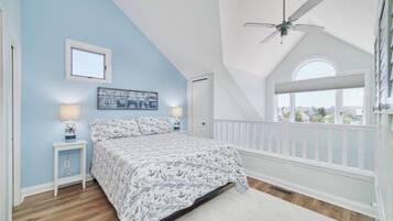 A bedroom with a sloped ceiling, a bed with patterned bedding, a nightstand with a lamp, a window with a view, and a ceiling fan.