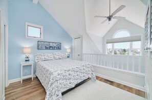 A bedroom with a sloped ceiling, a bed with patterned bedding, a nightstand with a lamp, a window with a view, and a ceiling fan. - Cozy 1-bedroom Pet Friendly Condo in Manistee.   Pool, Hot tub, 5 min to Beach! (Manistee)