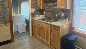 Una cocina con armarios de madera, pared de piedra, ventana con persianas y un cubo de basura.