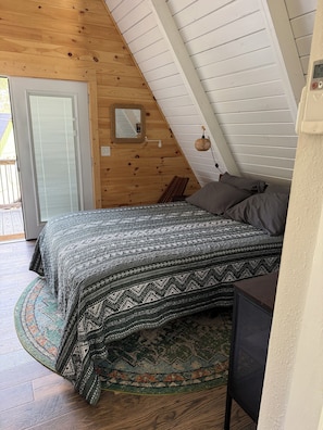 A bedroom with a bed, pillows, a small table, a wall-mounted light, and a door.