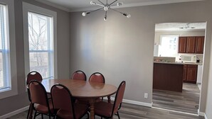 Uma sala de jantar com uma mesa redonda e seis cadeiras, um lustre no teto e uma cozinha ao fundo.