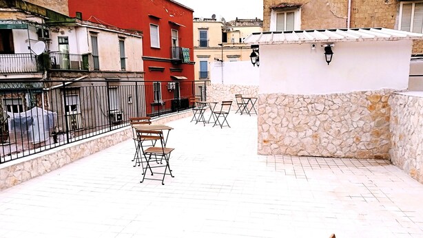 A narrow alleyway with a red building on the left and a white building on the right. There are outdoor seating areas with tables and chairs. - Terrazza Cristallini Napoli (Naples)