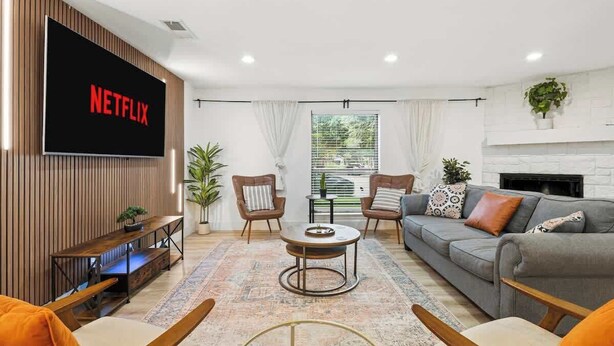 Ein modernes Wohnzimmer mit einem Flachbildfernseher, der 'NETFLIX' anzeigt, einem Sofa, zwei Sesseln, einem Couchtisch und einem Kamin.