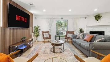 Un soggiorno moderno con una TV a schermo piatto che mostra 'NETFLIX', un divano, due poltrone, un tavolino da caffè e un camino.