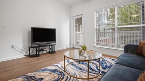 A living room with a flat-screen TV, a glass coffee table, a blue sofa, and large windows with blinds. - Chic Blue Apt w Patio + Pool, Free Parking, 10 min to ATL (Atlanta)