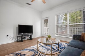 Ein Wohnzimmer mit Flachbildfernseher, einem Glastisch, einem blauen Sofa und großen Fenstern mit Jalousien.
